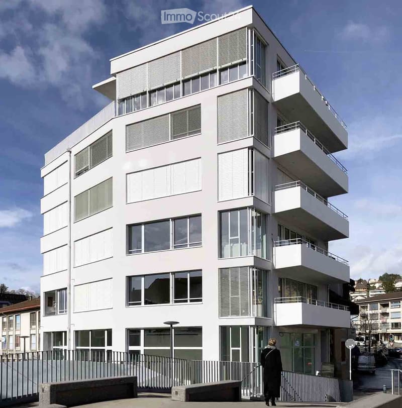 Modern white multi-story building with large windows and balconies, featuring contemporary architectural design.