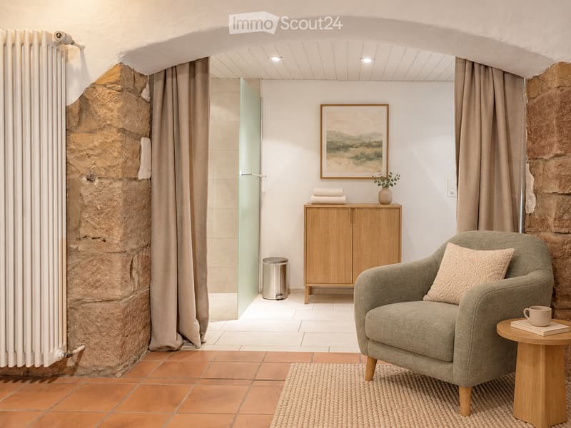 Cozy, well-lit room with a mix of modern and rustic elements. Features a comfortable armchair with a side table holding a cup and book, a wooden cabinet with a vase and folded towels, a framed picture on the wall, and a glass shower enclosure visible thro