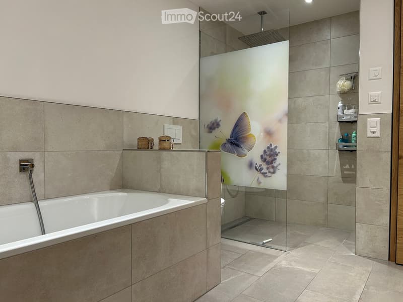 Modern bathroom with a bathtub, glass-enclosed shower with a decorative design, tiled walls, and shelves holding toiletries.