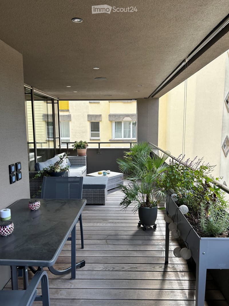 Balcony with wooden decking, modern furniture including a sofa, table, chairs, and several potted plants, with a view of neighboring buildings.