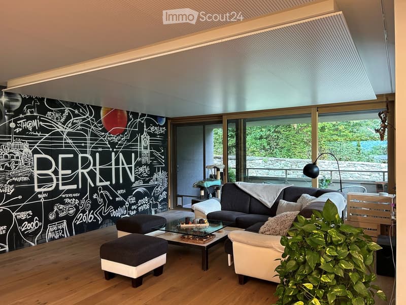 Modern living room with wooden flooring, large windows, a black wall with a 'BERLIN' mural, a dark grey sofa, a glass coffee table, ottomans, an armchair, and a floor lamp.