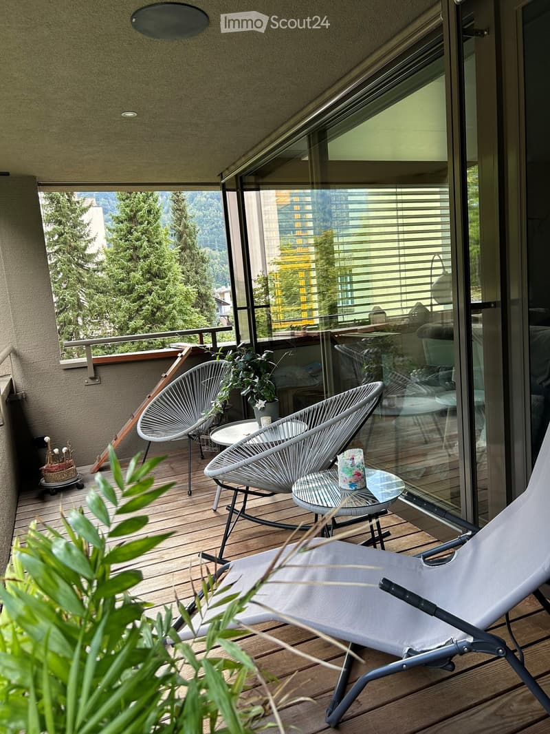 Balcony with wooden decking, modern chairs, small table, and potted plants, overlooking a cityscape with trees and buildings.