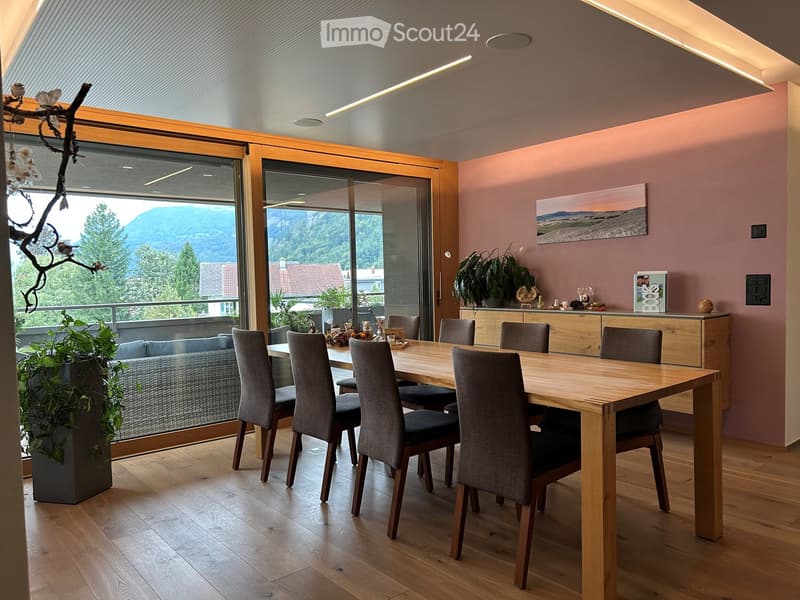Modern dining area with a wooden dining table, six chairs, a grey sofa, and a large window offering a scenic mountain view. The room features light pink walls, wooden flooring, and decorative plants.