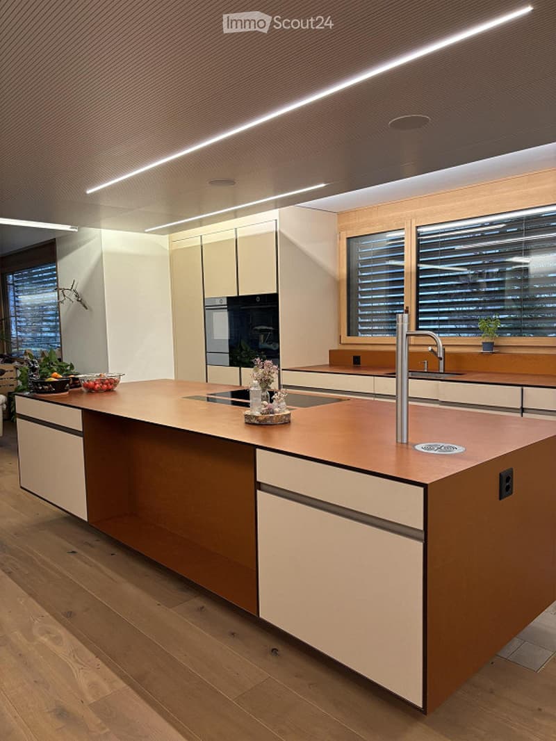 Modern kitchen with a central island, integrated sink, and sleek cabinetry. The space features wooden flooring, large windows with blinds, and built-in appliances including a fridge and oven.