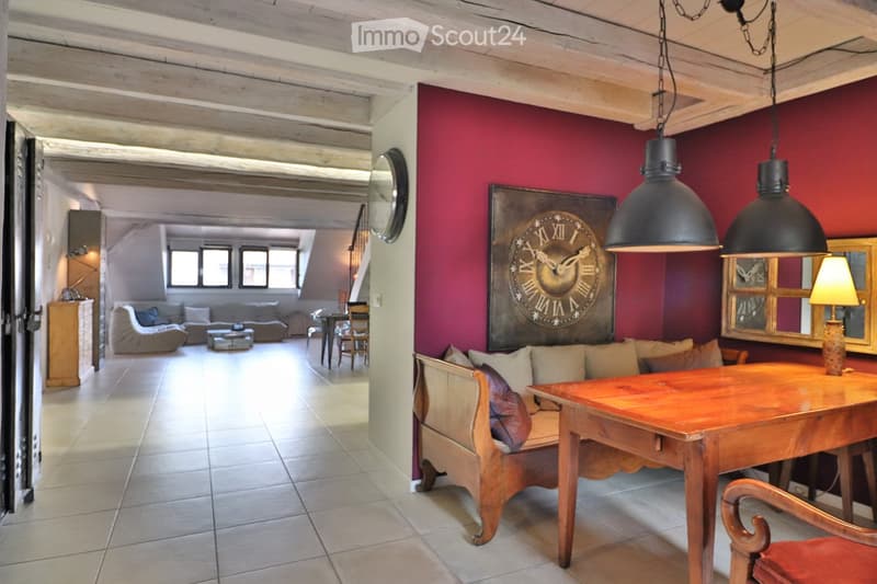 A spacious, well-lit living room with exposed wooden beams, a large red accent wall, and a mix of modern and rustic furniture including a sofa, bench, table, and chairs.