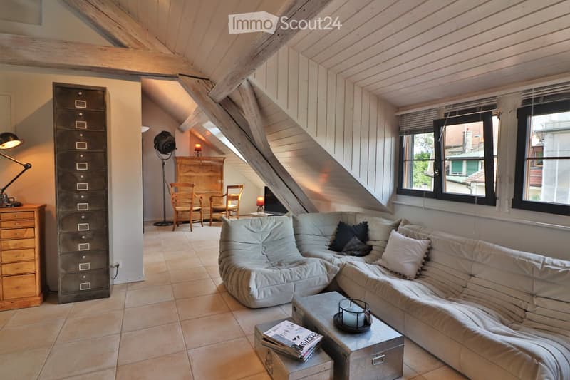 Cozy living room with exposed wooden beams, a large sectional sofa, a coffee table, two wooden chairs, a small table with a lamp, and a cabinet with drawers.