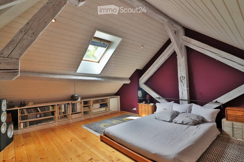 Cozy attic bedroom with wooden beams, slanted ceilings, skylight windows, and built-in bookshelf. Features a large bed with grey bedding, a wooden nightstand, and a speaker system.