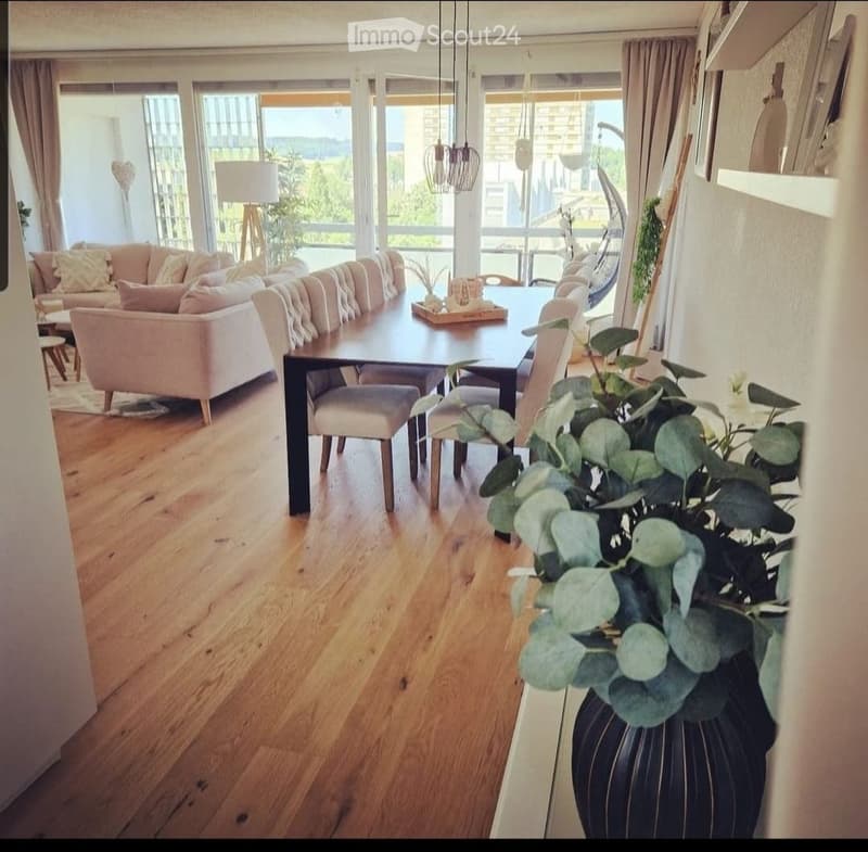 Open-plan living and dining area with wooden flooring, large windows offering a scenic view, a cream sofa, a dark wooden dining table with chairs, and a decorative flower vase on a side table.