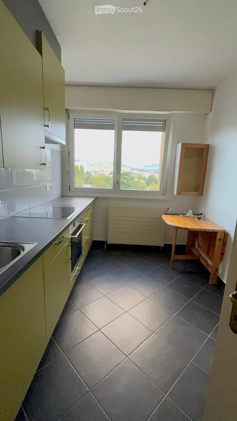 Empty kitchen with tiled floor, light-colored cabinets, sink, and a small wooden table near the window offering a view of the city and sea.