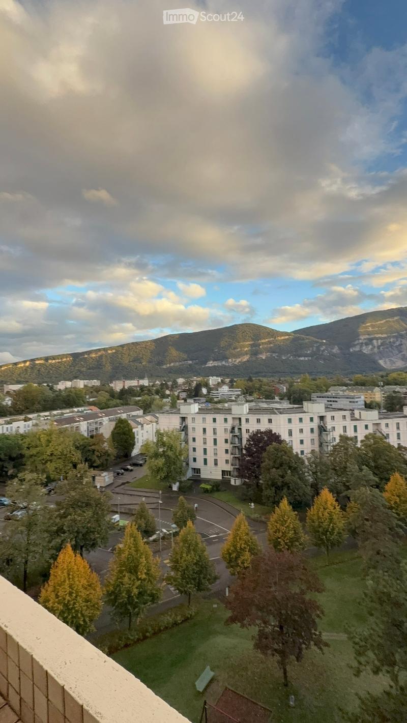 View from a balcony showing a cityscape with buildings, trees, a parking lot, and distant mountains under a partly cloudy sky.
