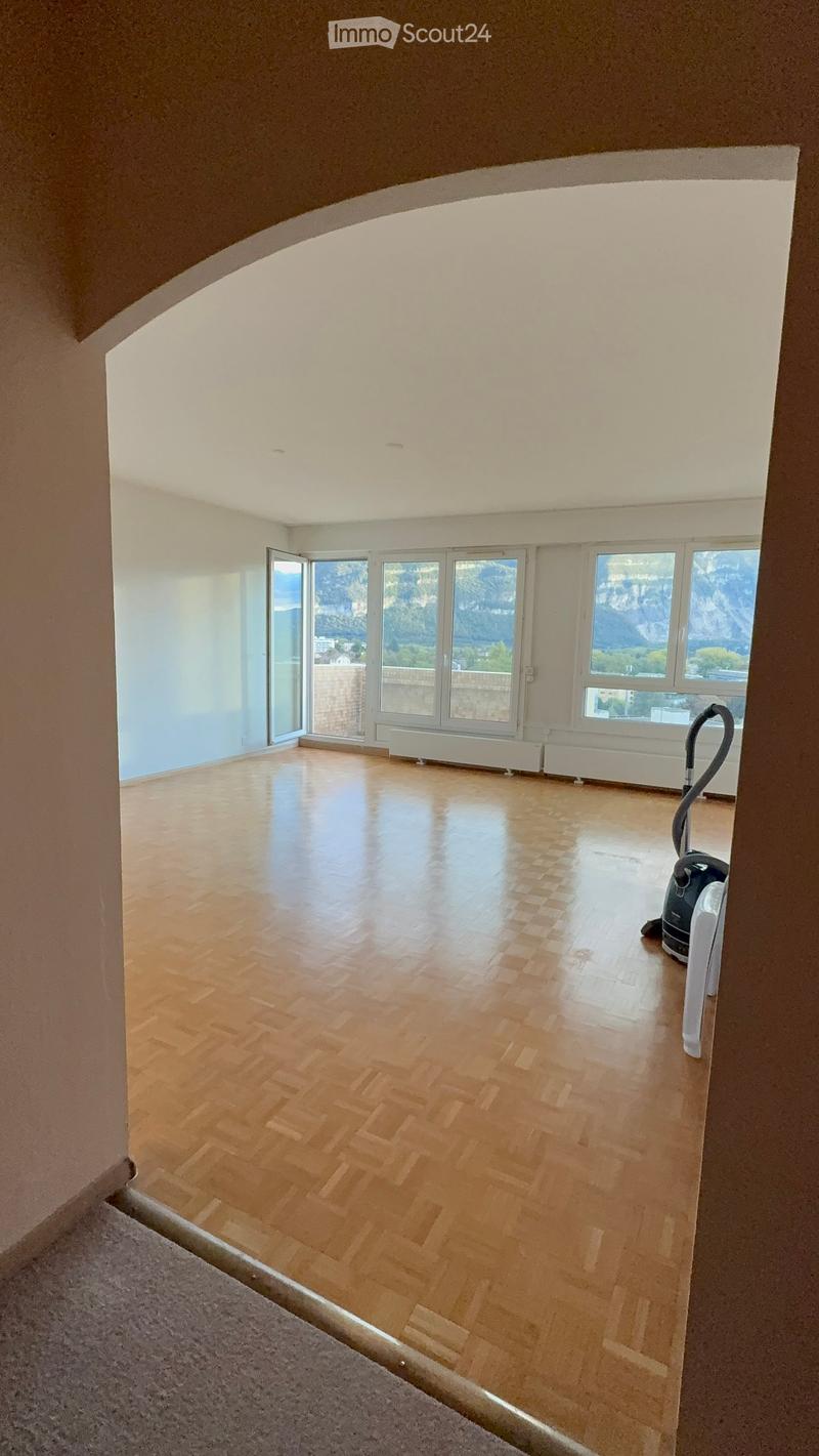 Empty room with wooden flooring, large windows offering a scenic mountain view, and a vacuum cleaner in the corner.