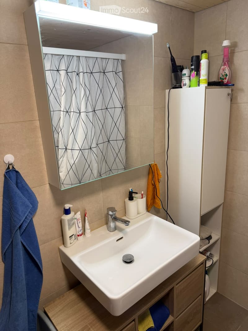 Modern bathroom with a white sink, mirror with built-in lighting, and various toiletries on the counter. Features a geometric-patterned shower curtain and a towel hanging on the side.