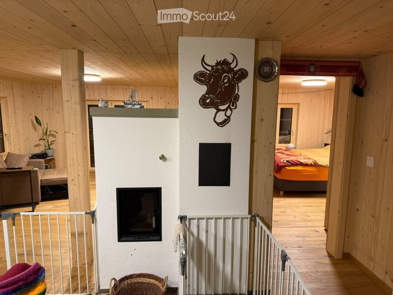 Cozy living room with wooden walls and ceiling, fireplace, sofa, small table with plant, and a visible bedroom with a bed in the background.