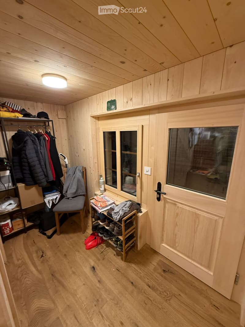 Cozy wooden-paneled room with a chair, shelf, small table, and various personal items including clothing and shoes.