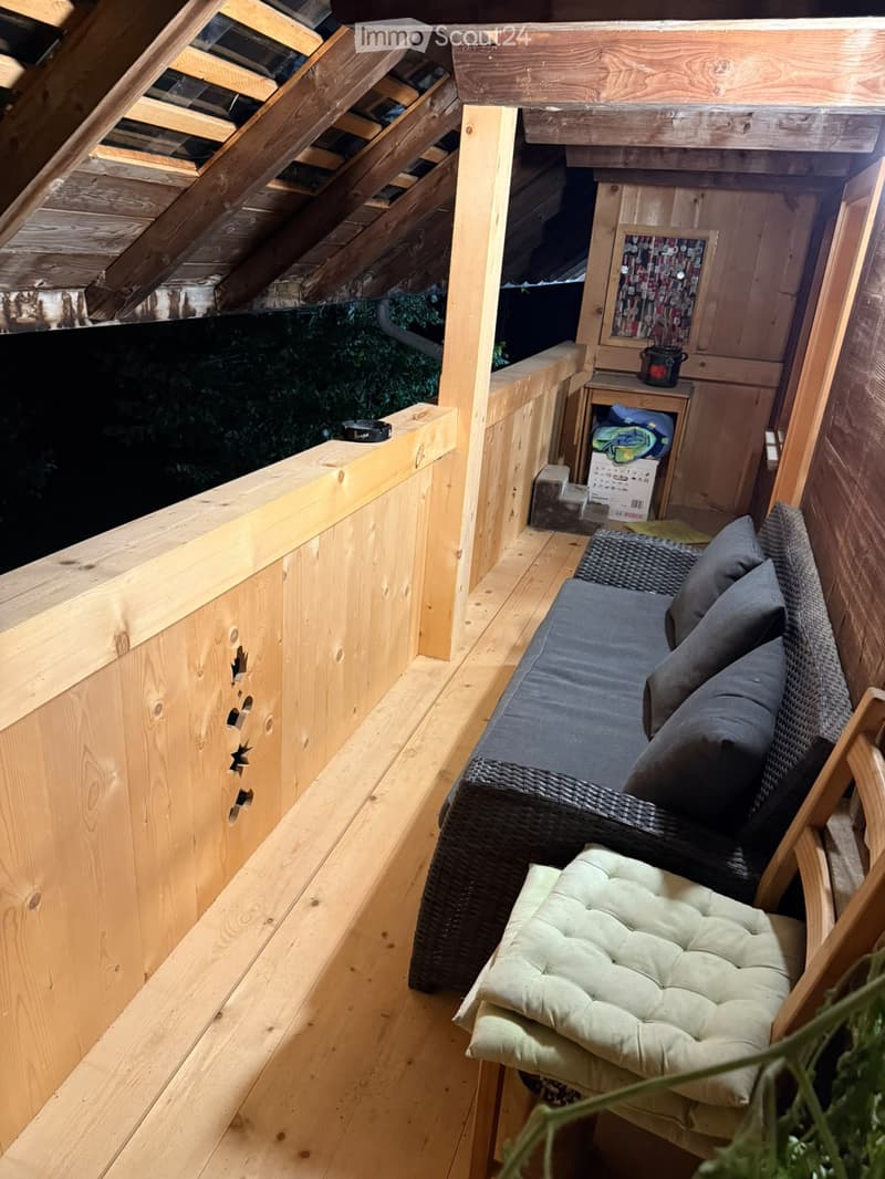 Wooden balcony with a wicker sofa, cushion, and small wooden table with items on it.