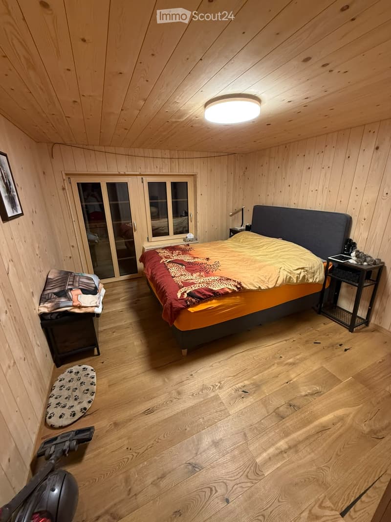 Cozy bedroom with wooden walls and floors, featuring a double bed with a colorful blanket, two nightstands with lamps, a picture frame on the wall, and a small stool with a patterned cushion.