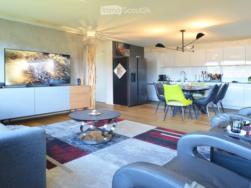 Modern living and dining area with a sofa, coffee table, dining table with chairs, kitchen area with cabinets and appliances, and a large TV on a stand.
