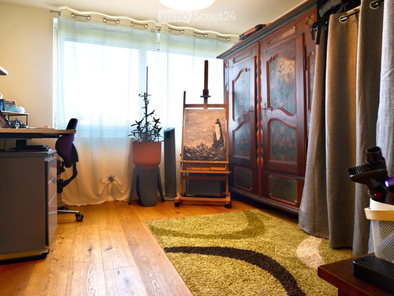 Well-lit room with wooden flooring, large window with sheer curtains, a potted plant, an easel with a painting, a wooden cabinet, a desk with a chair, and a small rug.