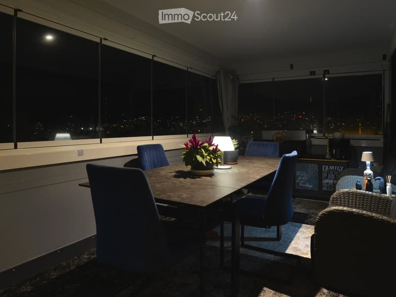 Modern dining area with a large wooden table, blue chairs, a potted plant, and a small lamp, overlooking a cityscape at night.