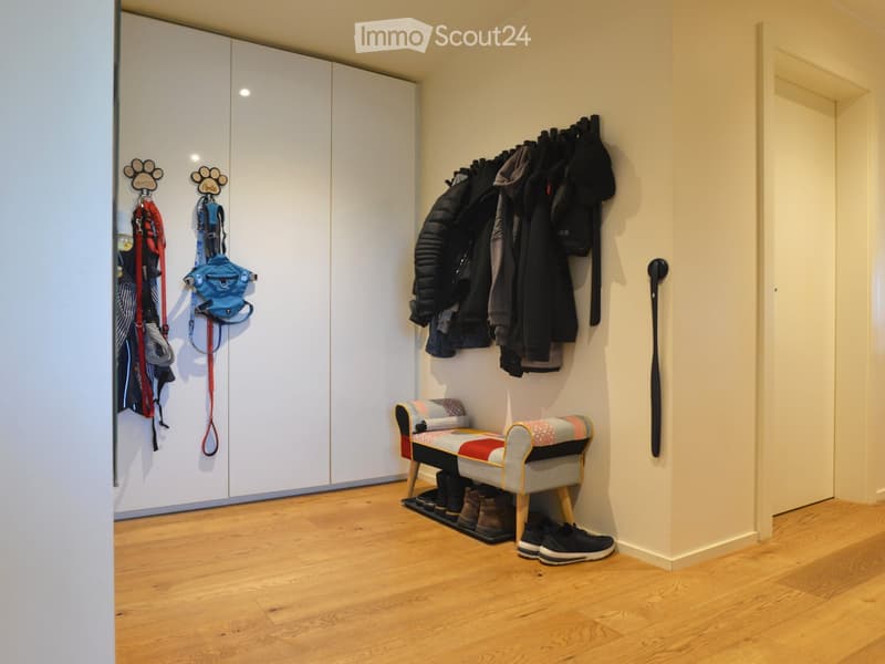 A well-lit hallway with wooden flooring, white closet doors, a bench with a colorful cushion, and various items including coats, shoes, and pet accessories.