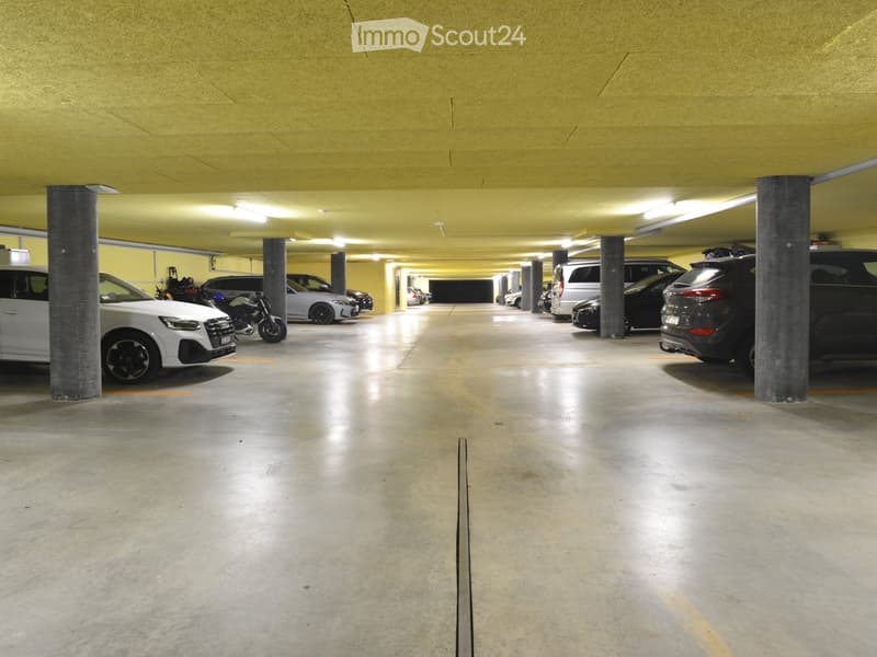 Underground parking area with several parked cars, motorcycles, and support pillars. The space is well-lit with a concrete floor and yellow-painted walls.