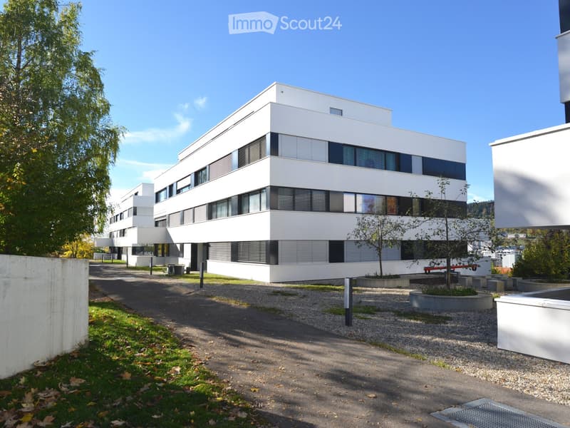 Modern, multi-story white building with large windows and a clean, minimalist design, surrounded by a gravel pathway and small landscaped areas.