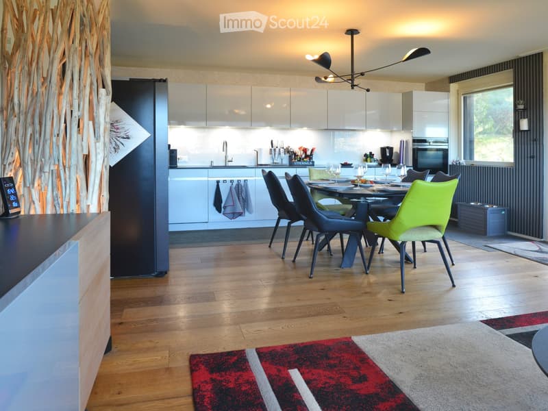 Modern kitchen and dining area with light-colored cabinets, a central island, and a dining table surrounded by colorful chairs. The space features wooden flooring, a red and white area rug, and contemporary lighting.