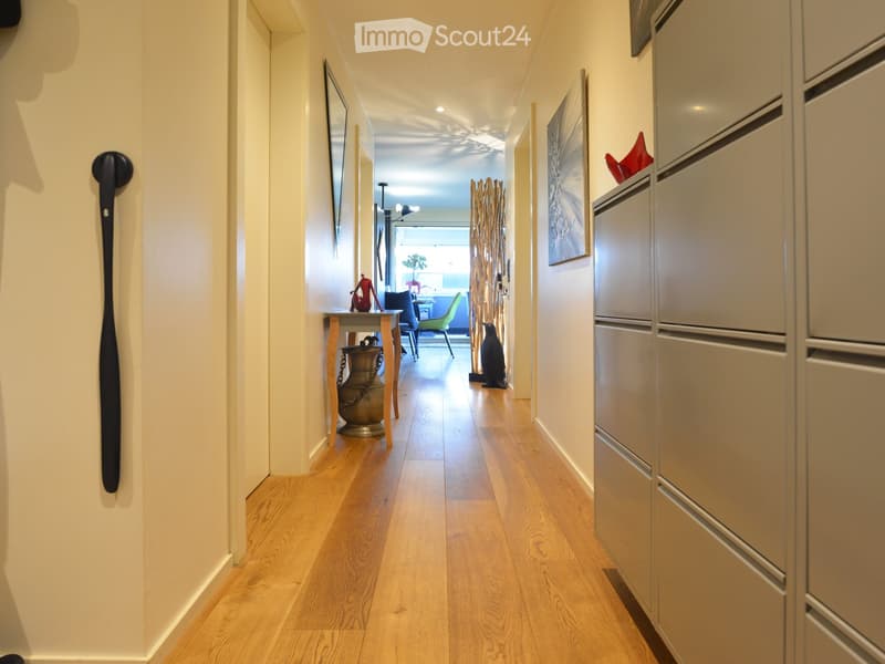 A well-lit hallway with light wooden flooring, white walls, and modern decor including a small table with a vase, chairs, and artwork.