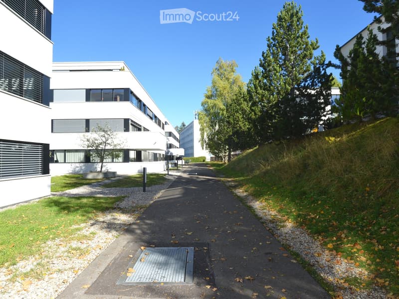 Modern white building with large windows and a raised ground floor, surrounded by greenery and a paved pathway.