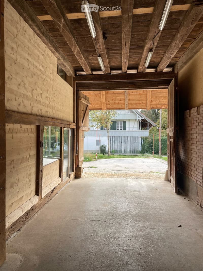 A long, open wooden corridor with exposed beams and brick walls, leading to an outdoor area with a paved ground and distant buildings.