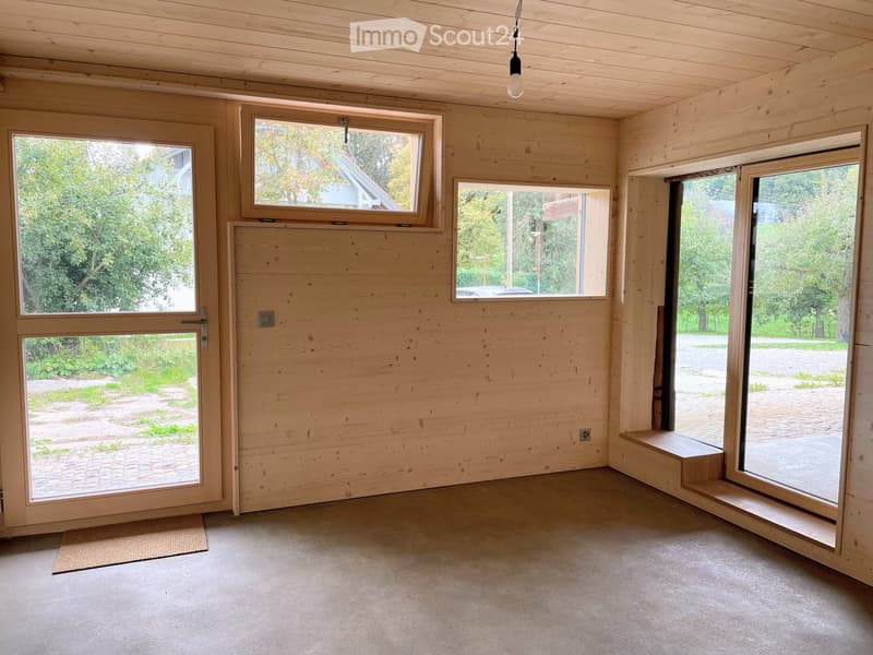 Empty room with wooden walls, large windows, and a door leading outside. The space has a simple, natural design with a light-colored floor and a small rug near the entrance.