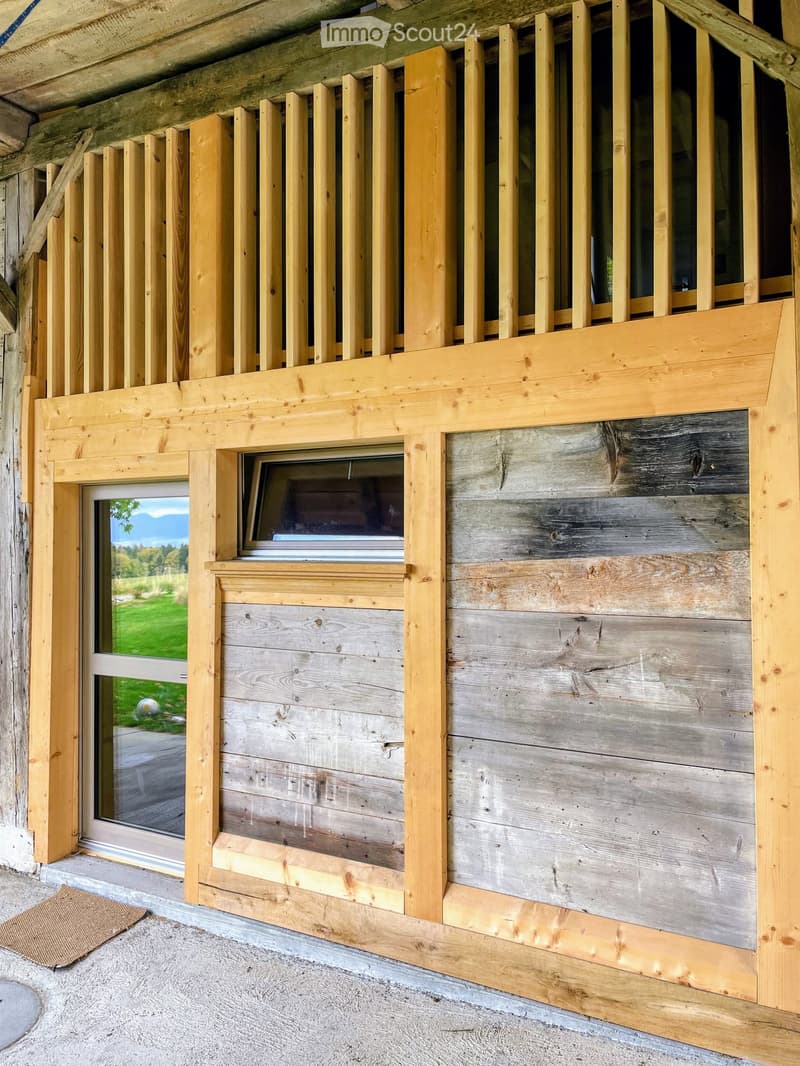 Wooden balcony with vertical slats, large window, and view of green landscape.