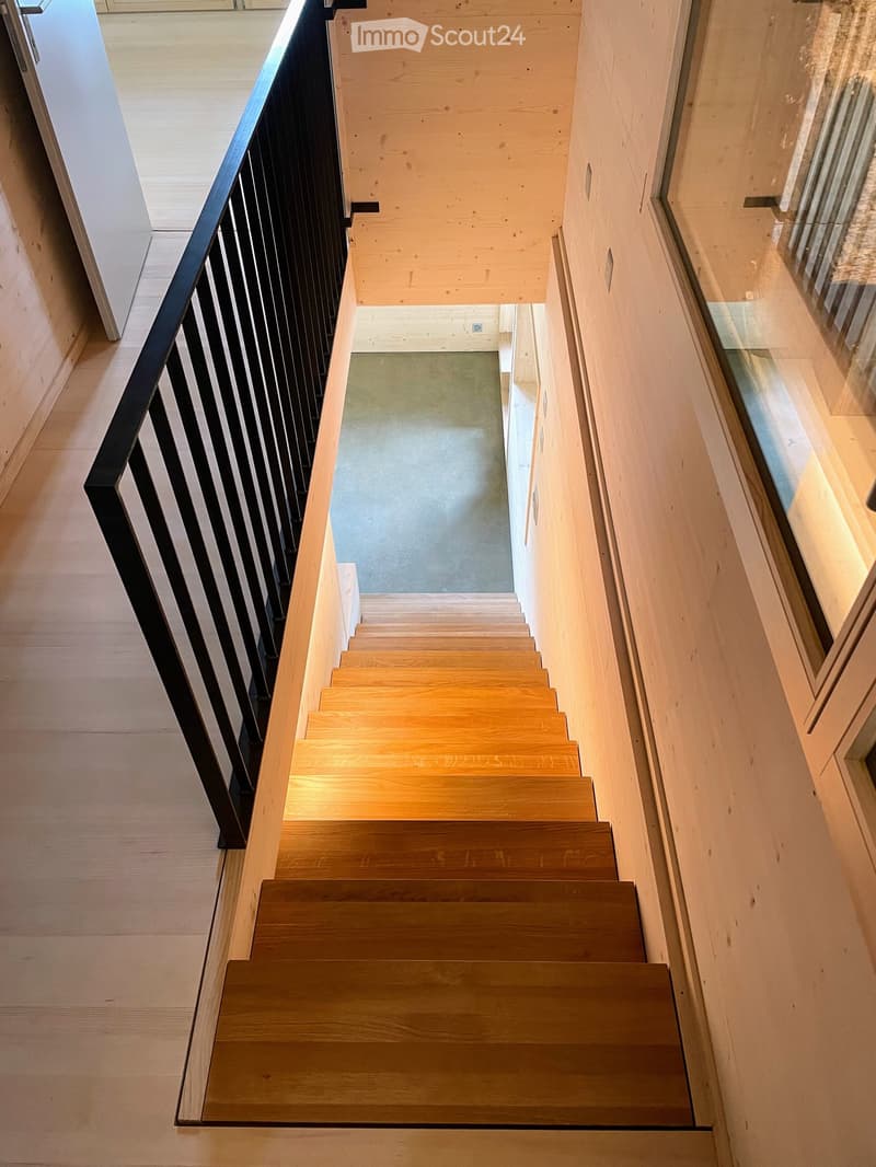 Wooden staircase with a black railing, light-colored wooden steps, and a glass side wall, leading to an upper level with a visible landing and railing.