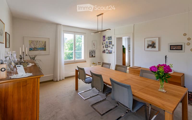 Bright dining room with a long wooden dining table surrounded by modern chairs, a vase with purple flowers, and a candle holder on a sideboard. The room features large windows with white curtains, framed artwork on the walls, and a cozy ambiance.