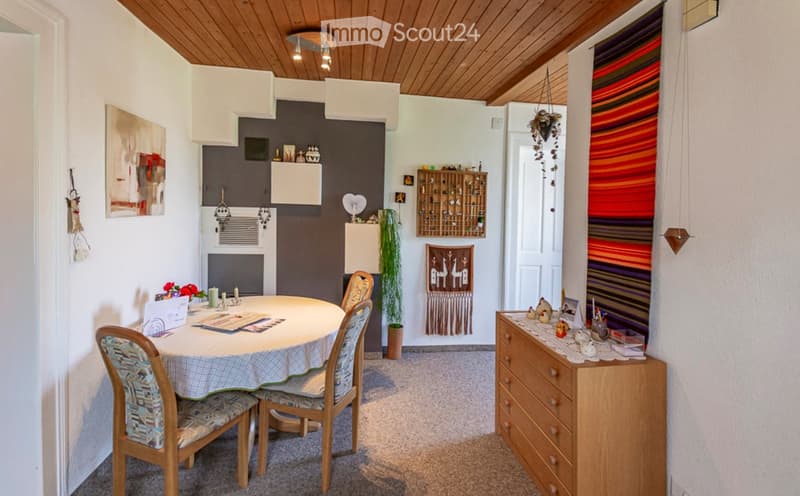 Dining area with a round table, four chairs, a wooden cabinet, and decorative items including a wall hanging and a plant. The room features a wooden ceiling and a colorful striped rug.
