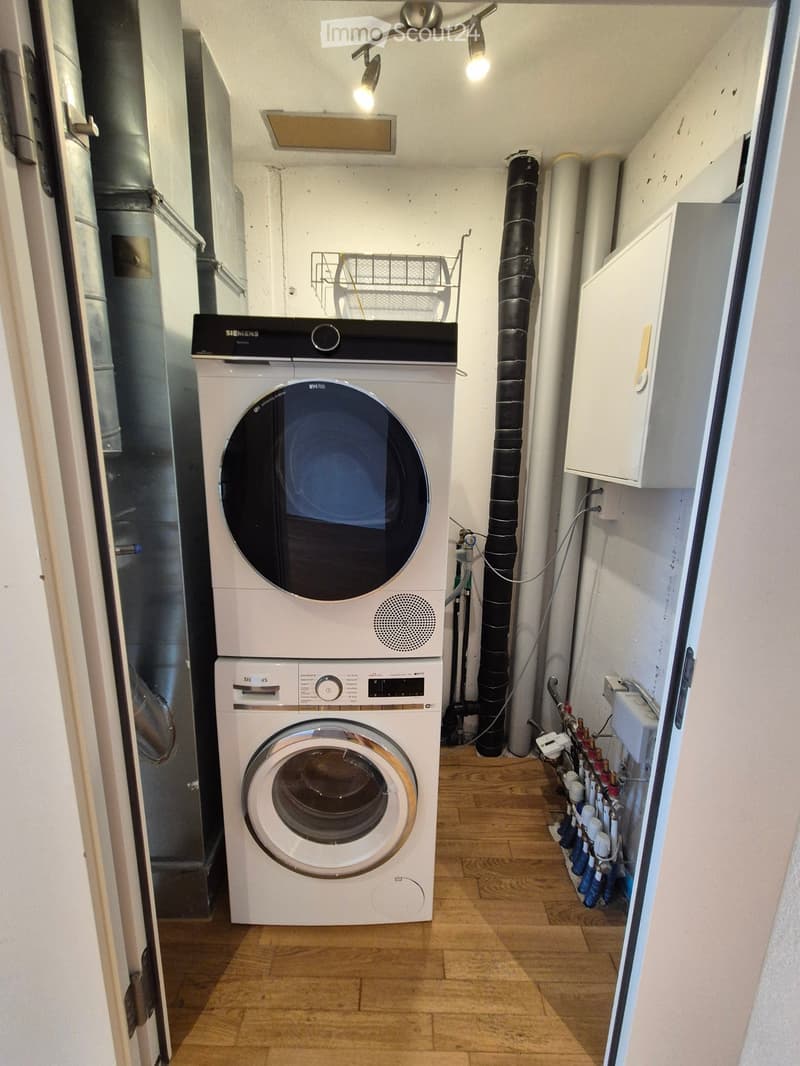 A small, well-lit room with wooden flooring, featuring a white washing machine and a white dishwasher stacked vertically, along with visible pipes and a small shelf.