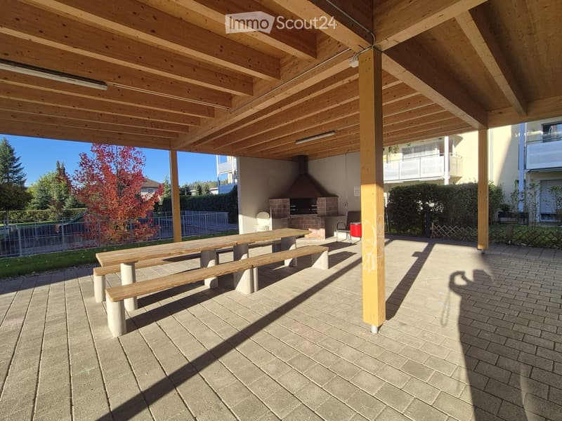 Wooden covered outdoor area with a brick barbecue, wooden tables and benches, surrounded by greenery and neighboring buildings.