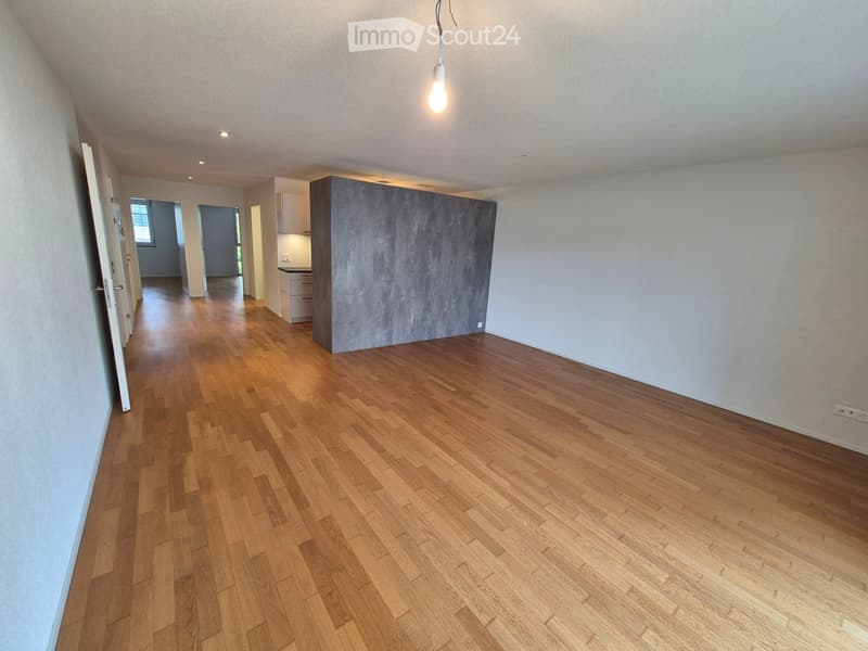 Empty room with wooden flooring, white walls, and a hanging light fixture. The room has open doors leading to other areas, including a kitchen with cabinets and a countertop.