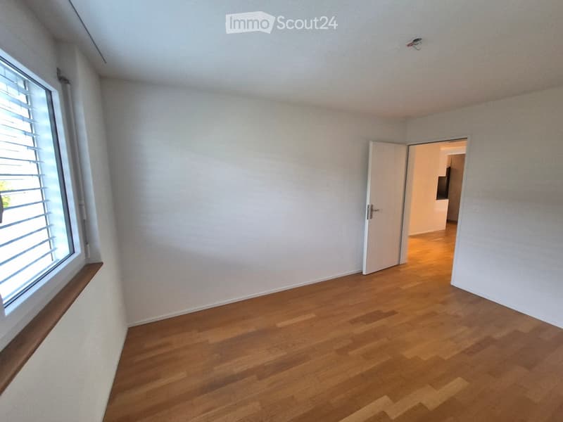 Empty room with wooden flooring, white walls, and a window with blinds. An open door leads to another room.
