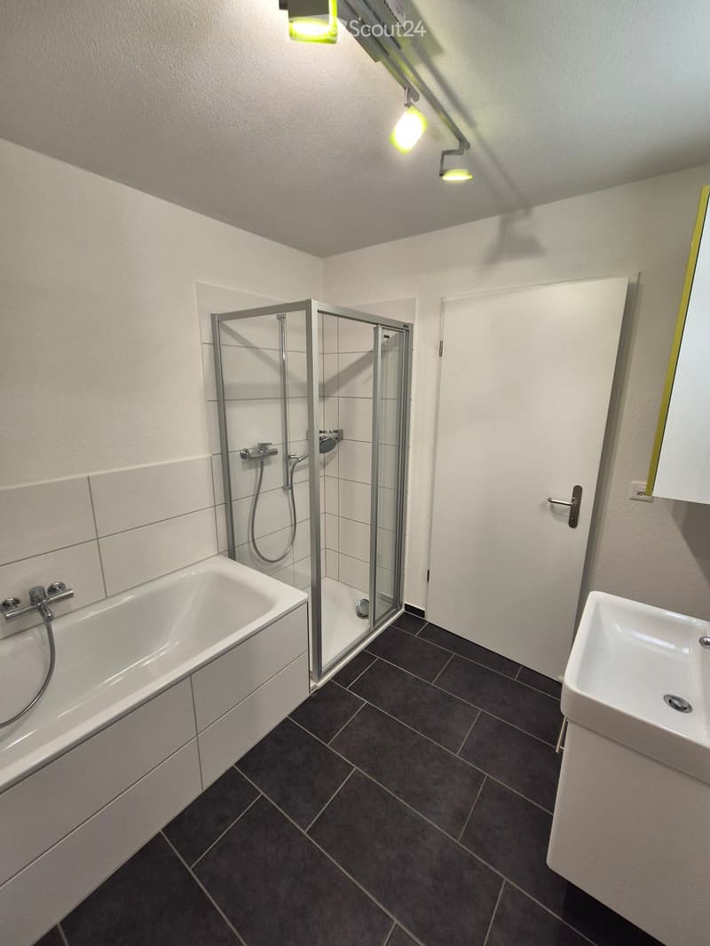 Modern bathroom with a bathtub, sink, and glass-enclosed shower stall. Features dark tiled flooring and white walls.