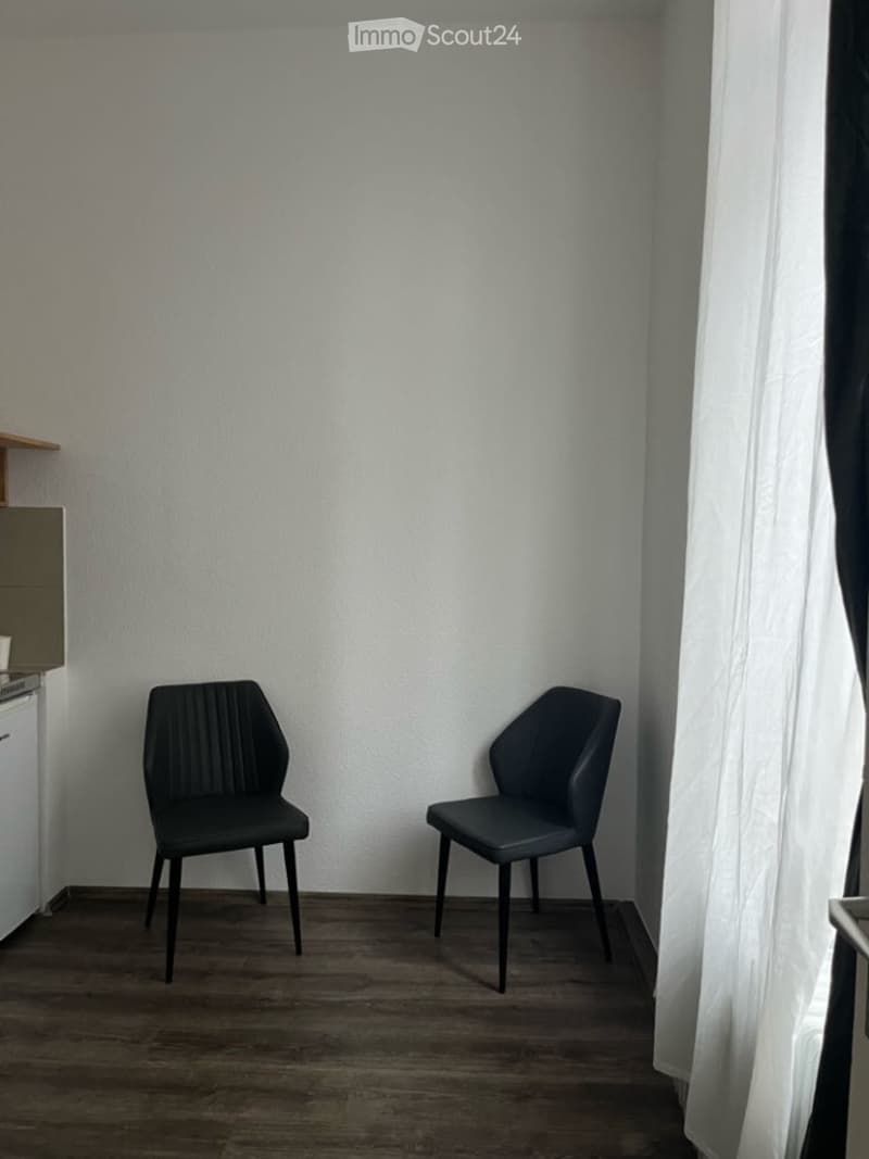 A small, unfurnished room with two modern black chairs, a wooden floor, and white walls. A small cabinet and a window with white curtains are visible.