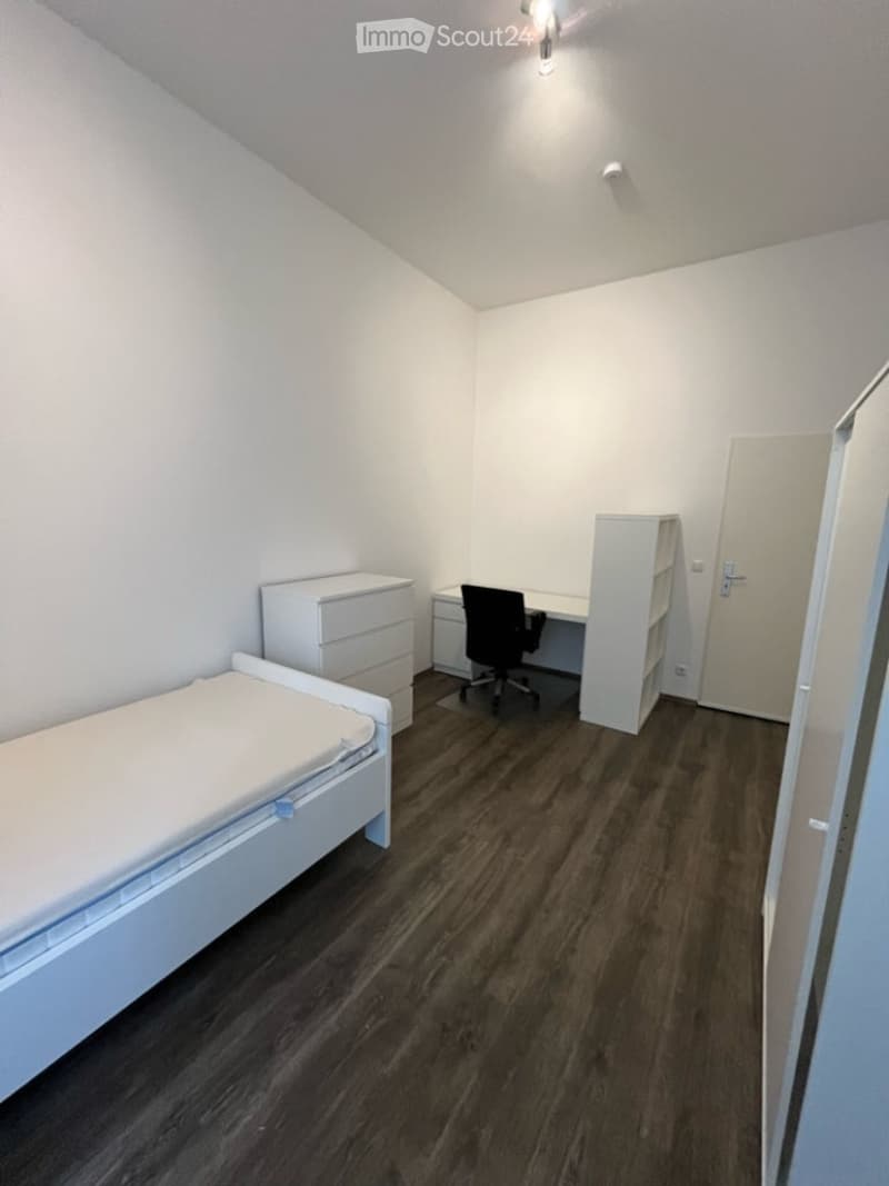 A small, modern bedroom with a single bed, a desk with a chair, a drawer unit, and a shelf. The room features wooden flooring and white walls.