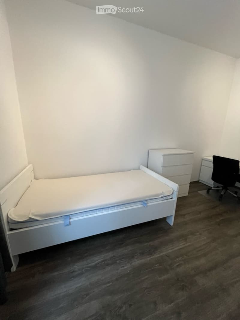 A small, neatly furnished bedroom with a single bed, a nightstand, a chair, and a desk. The room features light-colored walls and dark wooden flooring.