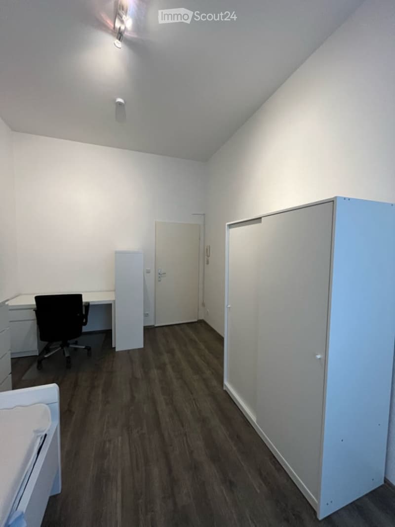 A small, furnished room with a wooden floor, white walls, and a slanted ceiling. It features a desk with a chair, a tall cabinet, and a door leading to another room.
