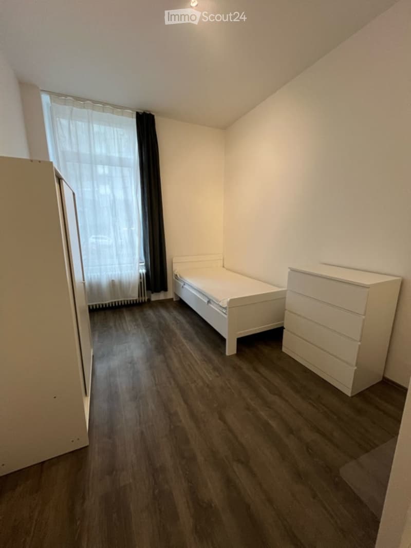 A small, furnished bedroom with a single bed, a white drawer unit, and a wardrobe. The room features wooden flooring and a window with white and black curtains.