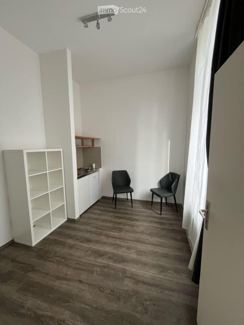 A modern, minimalist room with wooden flooring, white walls, and a small kitchen area. Two black chairs are placed near the kitchen counter, and a white shelving unit is visible on the left side.