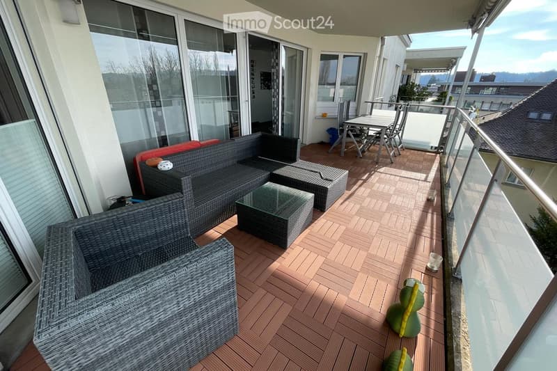Balcony with wooden decking, wicker sofa, glass coffee table, dining table and chairs, and a view of the surrounding area.