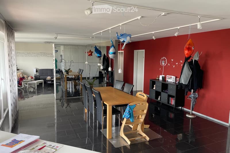 Open-plan living and dining area with a red accent wall, a wooden dining table with chairs, a sofa, a small cabinet, and decorative balloons.
