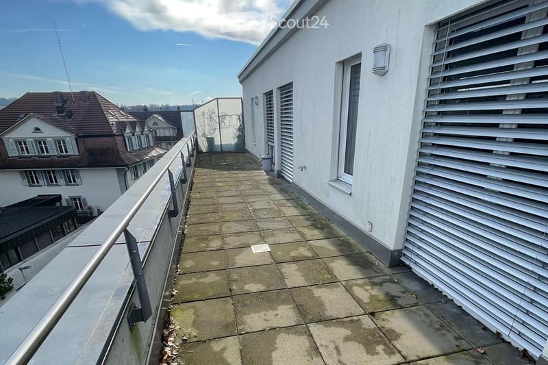Balcony with tiled floor, metal railing, and white exterior walls. The view shows neighboring houses and a clear sky.