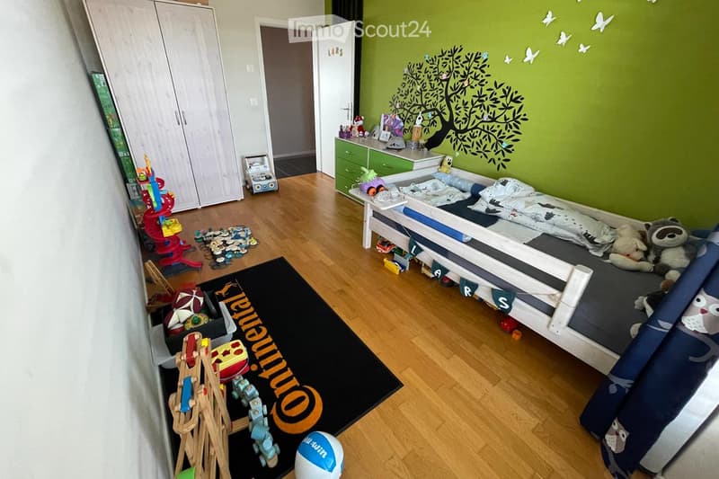 A well-lit children's bedroom with a single bed, colorful toys, a wardrobe, and a green wall with a tree mural.